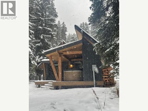 Snowy rear exterior with covered deck and stacked firewood at 1166 Apex Mountain Road, Penticton Apex